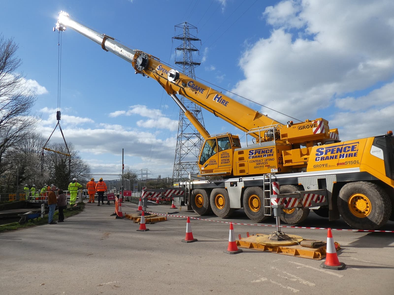 Mobile crane kuning dengan boom terentang mengangkat beban di area kerja yang dipasangi kerucut pengaman